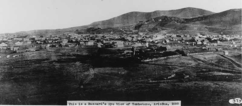 Historic black and white photograph showing a bird's-eye view of Tombstone, Arizona in 1880. The image captures a sprawling frontier town nestled in a desert valley, with numerous small buildings scattered across the landscape. In the background, rugged hills and mountains can be seen. The town appears to be in its early stages of development, with open spaces between clusters of structures. A caption at the bottom of the image reads 'This is a Buzzard's Eye View of Tombstone, Arizona, 1880'.